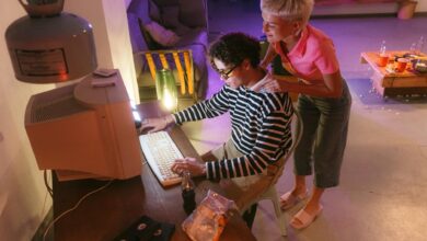 Two friends in a cozy room share a moment using a vintage desktop computer, illuminated softly.