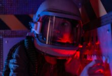 A woman astronaut wearing a spacesuit in a control room lit by red lights, looks focused.