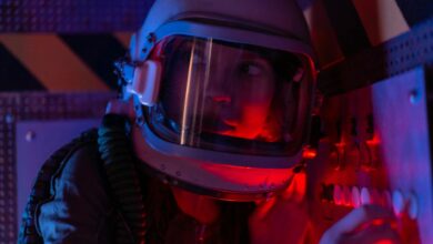 A woman astronaut wearing a spacesuit in a control room lit by red lights, looks focused.