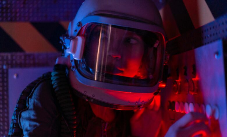 A woman astronaut wearing a spacesuit in a control room lit by red lights, looks focused.