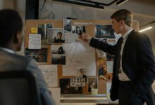 Two detectives discussing suspects and evidence on a cork board in an office setting.
