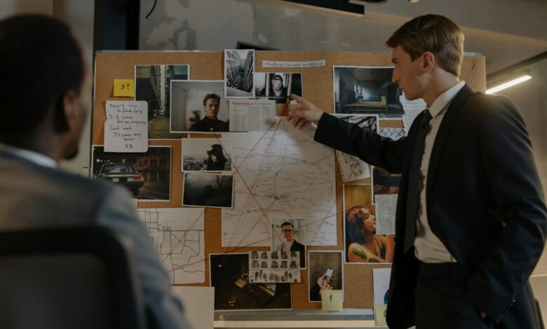 Two detectives discussing suspects and evidence on a cork board in an office setting.