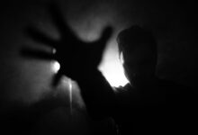 Black and white of anonymous male reaching out hand to camera while standing near wall with lamp in dark room