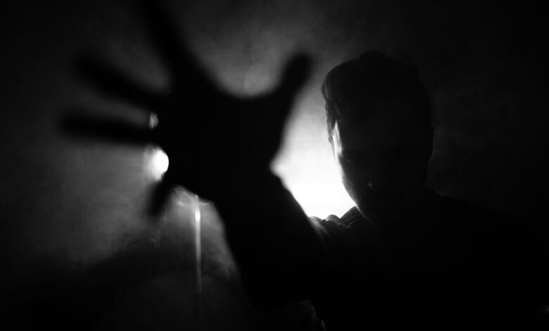 Black and white of anonymous male reaching out hand to camera while standing near wall with lamp in dark room