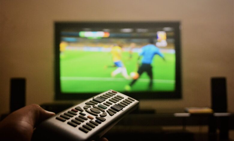 Close-up of a hand holding a remote control in front of a TV showing a football match.
