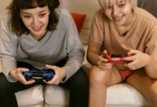 Low angle of cheerful crop Asian females in home clothes playing video games with joysticks in hands on sofa in living room