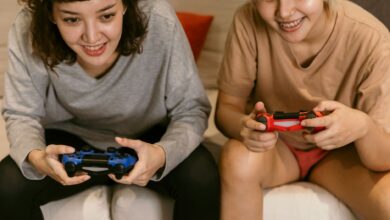 Low angle of cheerful crop Asian females in home clothes playing video games with joysticks in hands on sofa in living room