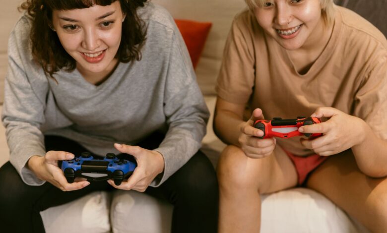 Low angle of cheerful crop Asian females in home clothes playing video games with joysticks in hands on sofa in living room