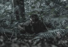 A man crouches in dense forest foliage, blending with his surroundings, evoking a sense of adventure and stealth.