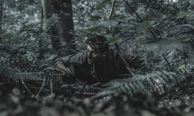A man crouches in dense forest foliage, blending with his surroundings, evoking a sense of adventure and stealth.