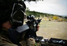 A soldier wearing camouflage and headphones aiming a rifle during outdoor target practice session.