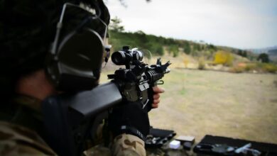 A soldier wearing camouflage and headphones aiming a rifle during outdoor target practice session.