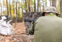 A man in camouflage gear aims during a paintball match in a forest setting.