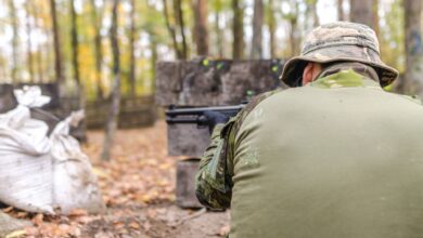 A man in camouflage gear aims during a paintball match in a forest setting.