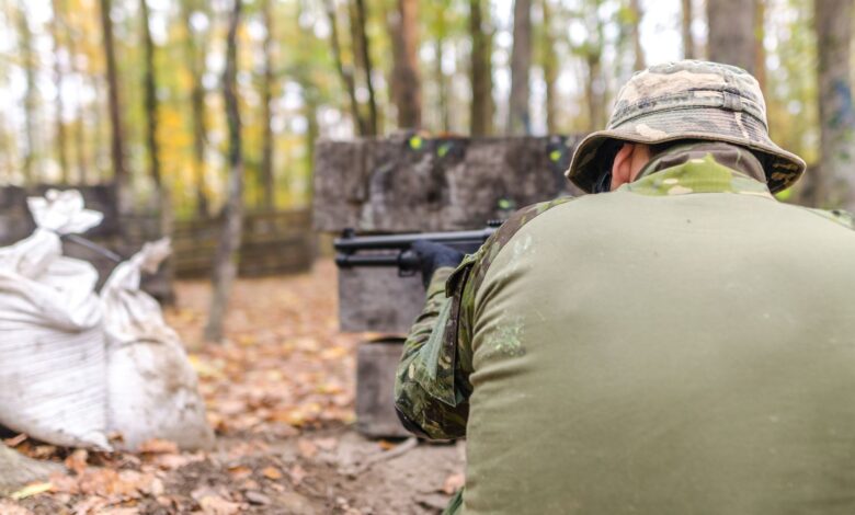A man in camouflage gear aims during a paintball match in a forest setting.