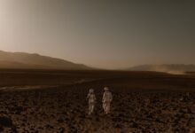 Two astronauts in spacesuits walk across a Mars-like desert landscape during the day.