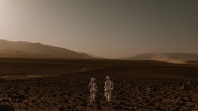 Two astronauts in spacesuits walk across a Mars-like desert landscape during the day.