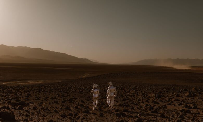 Two astronauts in spacesuits walk across a Mars-like desert landscape during the day.