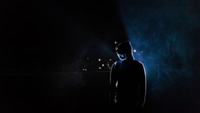 Silhouette of a man in a glowing LED mask and hoodie in a dark, foggy setting.