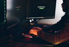 Side view of crop anonymous male cyber thief accessing information on desktop computer screens at dusk