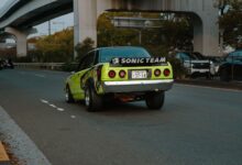 A vintage green car drives on an urban highway in Tokyo with distinct banners and decals.