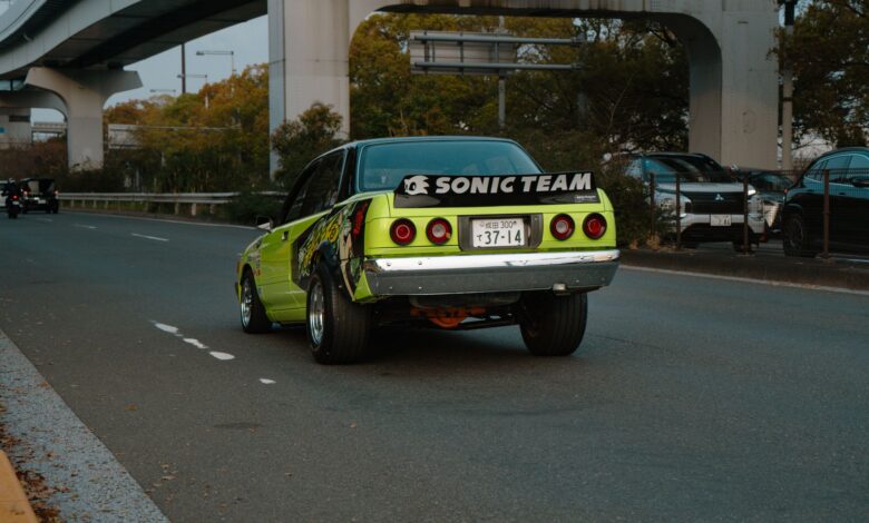 A vintage green car drives on an urban highway in Tokyo with distinct banners and decals.