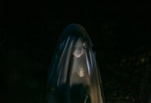 Mysterious little girl standing in darkness covered with white blanket as ghost and shining flashlight on face