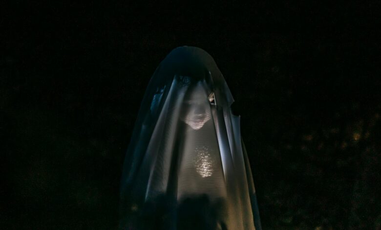 Mysterious little girl standing in darkness covered with white blanket as ghost and shining flashlight on face