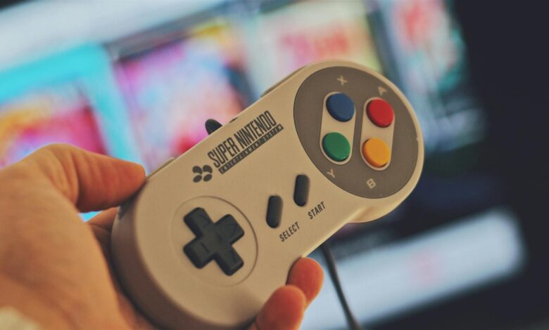 Hand holding a classic Super Nintendo game controller indoors with a blurred screen in the background.