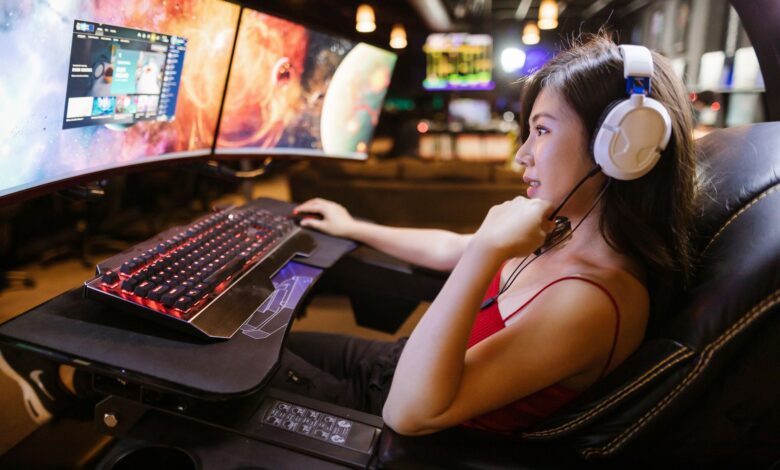 Young woman in red top gaming on dual monitors with white headphones in a modern setup.