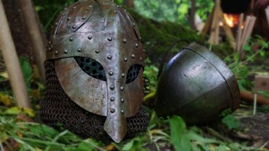 Close-up of a medieval helmet with chainmail on a forest floor, surrounded by leaves and natural elements.