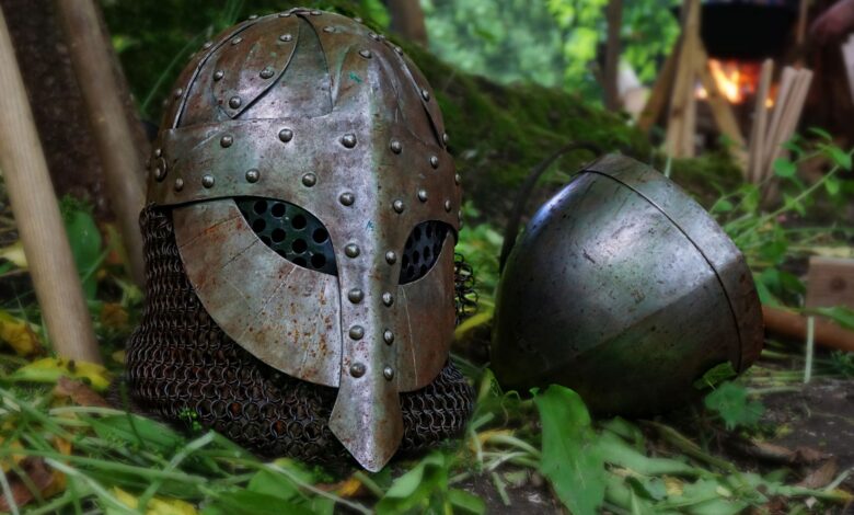 Close-up of a medieval helmet with chainmail on a forest floor, surrounded by leaves and natural elements.