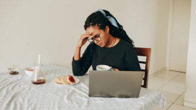 Woman working from home, stressed, drinking coffee, seated at table with laptop.