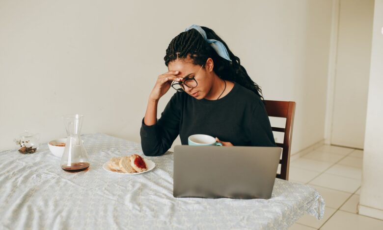 Woman working from home, stressed, drinking coffee, seated at table with laptop.
