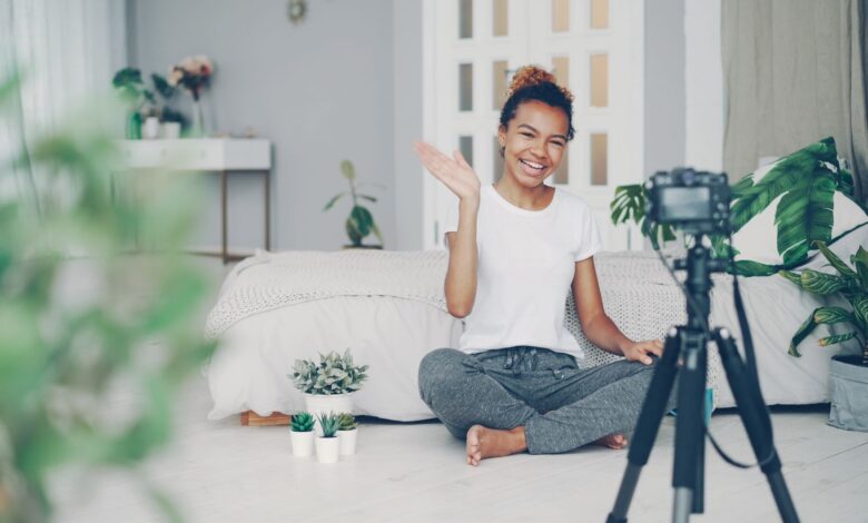 Smiling woman waves at camera while recording a vlog in a stylish bedroom setting.