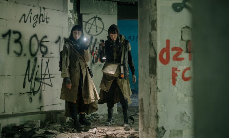 Two people exploring an abandoned, graffiti-filled building with flashlights, evoking a post-apocalyptic atmosphere.