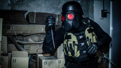 Individual in tactical gear and gas mask holding a weapon in an abandoned warehouse.