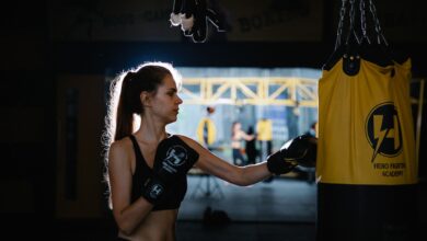 Serious fit young sportswoman with long hair in activewear and gloves practicing punches on hanging bag while working out in boxing club