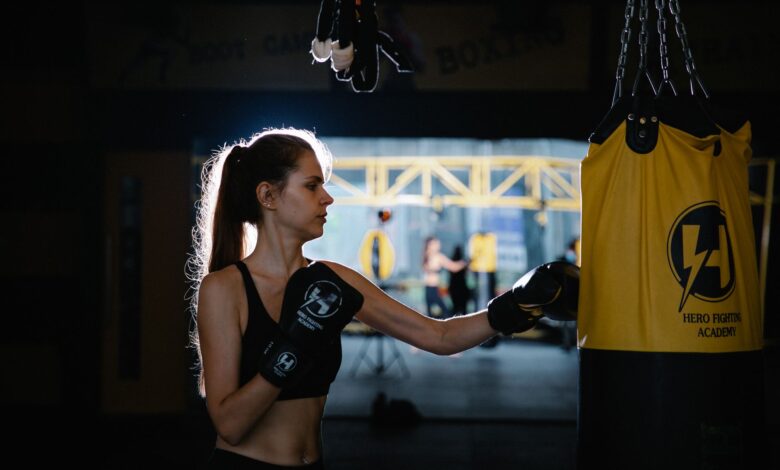 Serious fit young sportswoman with long hair in activewear and gloves practicing punches on hanging bag while working out in boxing club