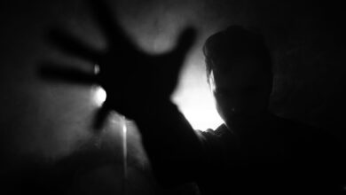 Black and white of anonymous male reaching out hand to camera while standing near wall with lamp in dark room