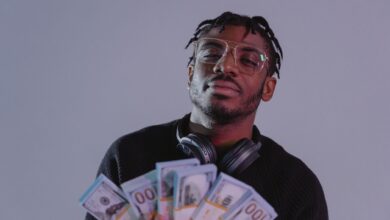 Portrait of a man holding bundles of cash wearing glasses and headphones. Indoor setting.