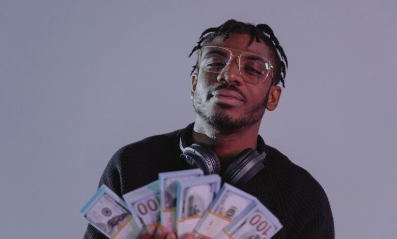 Portrait of a man holding bundles of cash wearing glasses and headphones. Indoor setting.