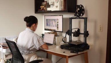 A woman working on a laptop with a 3D printer in a modern home office setup.