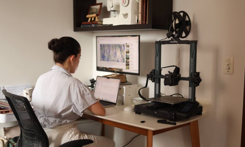 A woman working on a laptop with a 3D printer in a modern home office setup.