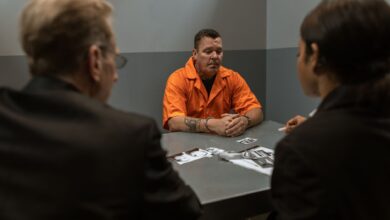 Prisoner in orange jumpsuit sits handcuffed during an interrogation with detectives in a room.