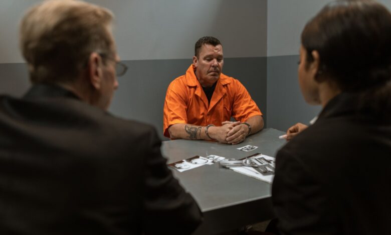 Prisoner in orange jumpsuit sits handcuffed during an interrogation with detectives in a room.