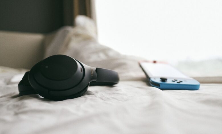 Close-up of a black headset and a gaming console resting on a soft, white bedspread in a calm, indoor setting.