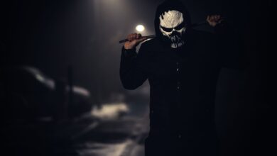 A spooky masked figure holding a weapon in a dimly lit street at night.