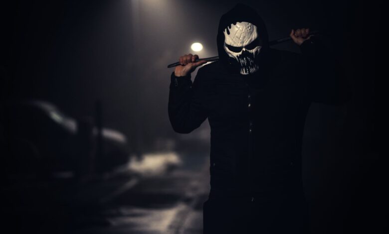 A spooky masked figure holding a weapon in a dimly lit street at night.