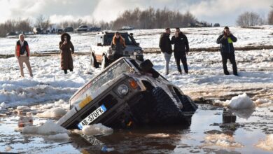 An off-road SUV is stuck in muddy snow with people standing nearby in Gjinari, Albania.